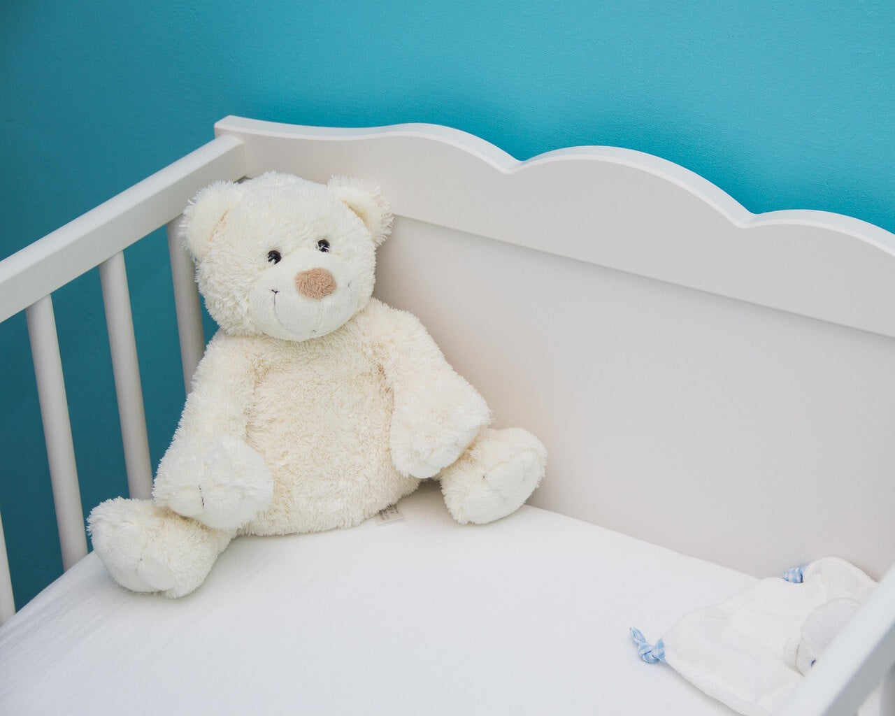 Teddybär sitzt in einem Babybett und neben an ist ein Kuscheltuch