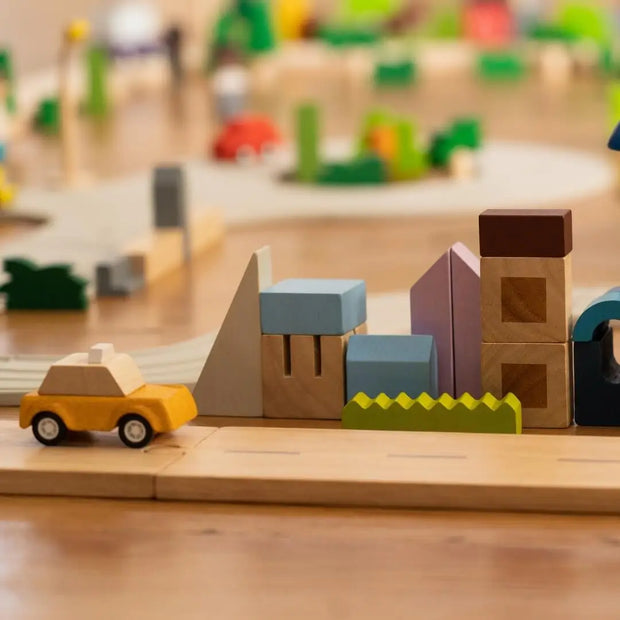 Holzbausteine in verschieden formen und großen im Stil einer Stadt aufgebaut, mit einer Plantoys Straße und einem gelben Holzauto darauf